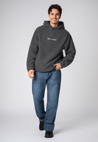 Grauer Fleece-Hoodie mit Kängurutasche und "SBLVL STUDIO"-Logo, kombiniert mit blauen Jeans und schwarzen Schuhen. Lässiges Design und entspannte Passform.