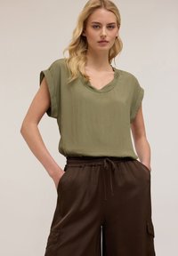 Blusa verde oliva comoda con maniche arrotolate, design a V, abbinata a pantaloni marroni con cordoncino e tasche laterali.