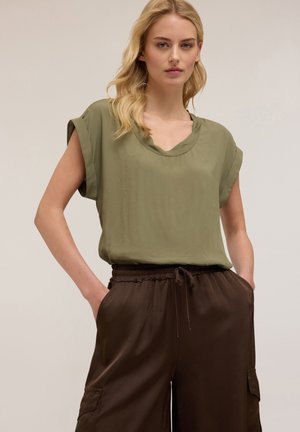 Blusa verde oliva comoda con maniche arrotolate, design a V, abbinata a pantaloni marroni con cordoncino e tasche laterali.