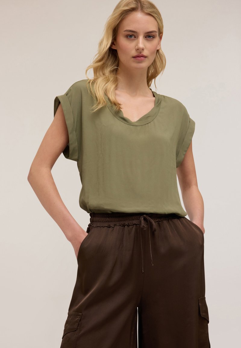 Blusa verde oliva comoda con maniche arrotolate, design a V, abbinata a pantaloni marroni con cordoncino e tasche laterali.
