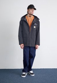Zwarte waterdichte jas met meerdere zakken, een ritsborzak en een verstelbare capuchon, gedragen over een oranje shirt en blauwe broek.
