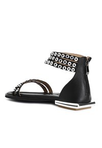 Musta sandaali, jossa on kolme leveää hihnaa, jotka on koristeltu metallisilla niiteillä, takana vetoketjukiinnitys ja neliömäinen geometrinen kantapäätekijä.