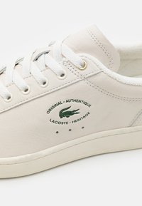 White leather sneaker with textured surface, round toe, metal eyelets, white laces, and a green logo patch. Sole is cream-colored and thick.