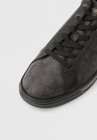 Μαύρο αθλητικό παπούτσι με υφή σ το πάνω μέρος, λείες δερμάτινες αντιθέσεις και μαύρα επίπεδα κορδόνια. Ορατή σόλα από καουτσούκ.