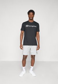 Schwarzes Baumwoll-T-Shirt mit weißem "Champion"-Logo, kombiniert mit grauen Shorts. Das Model trägt weiße Sneakers und Socken mit einem Logo.