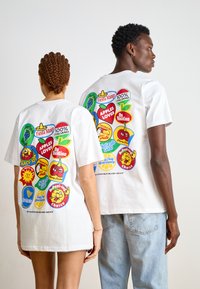 Hvite bomulls-t-skjorter har fargerike frukt-tema grafikker på baksiden, med ulike logoer og lekne design i lyse farger.
