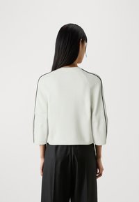 Maglione bianco a coste con maniche a tre quarti, caratterizzato da strisce nere laterali e un collo rotondo, indossato sopra pantaloni neri.