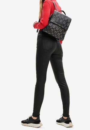 Žena v černých úzkých kalhotách, s černým květovaným batohem, v červeném dlouhém rukávu a černých teniskách, stojící před bílým pozadím.