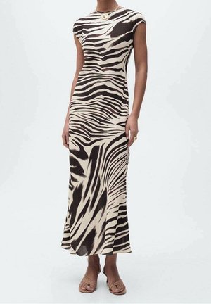 Kobieta ubrana w bezrękawnikową sukienkę o długości do kostek w zebra print, beżowe sandały i złoty naszyjnik z okrągłą zawieszką.