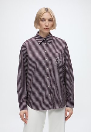 Jeune femme aux cheveux blonds courts porte une chemise rayée ample à manches longues boutonnée et un pantalon blanc devant un fond uni.
