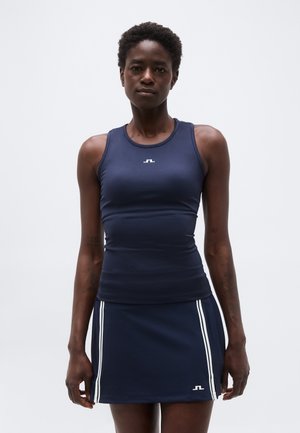 Γυναίκα που φοράει μπλε σκούρο αμάνικο τοπ και αντίστοιχη φούστα με λευκές κάθετες ρίγες και μικρό λογότυπο σε κάθε κομμάτι, στέκεται μπροστά σε απλό φόντο.