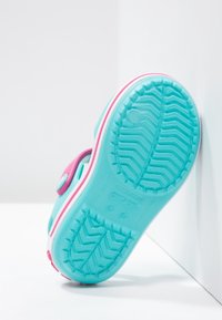 Sininen ja vaaleanpunainen lasten puutarhakenkä, jossa on teksturoitu pohja, nojaa valkoista seinää vasten valkoisella pinnalla.