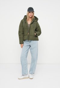 Olivgrüner Puffer-Jacke mit cropped Design und Kapuze, kombiniert mit hellblauen Jeans und weißen Sneakers. Eine dunkle Mütze rundet den Look ab.