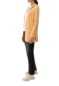 Cardigan arancione con cappuccio, maniche lunghe e spacchi laterali, abbinato a una maglietta bianca e jeans neri. Modella che indossa sneakers di colore chiaro.