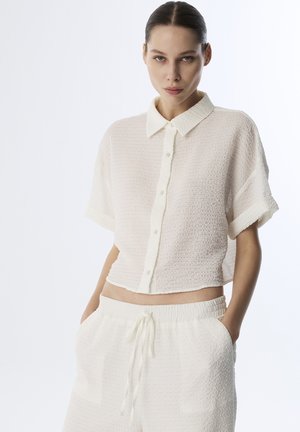 Witte cropped button-up shirt met een gestructureerde stof, korte mouwen en een zachte kraag, gecombineerd met elastische shorts in hetzelfde materiaal.