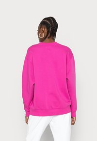 Persona con capelli intrecciati, che indossa orecchini a cerchio argentati e una felpa lunga a maniche con il collo alto di colore rosa acceso, con pantaloni bianchi, di spalle rispetto alla fotocamera.