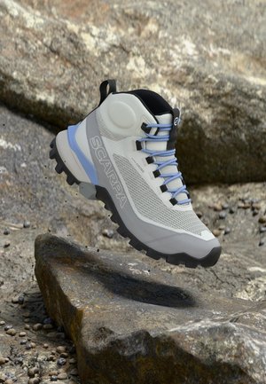 Helehall ja sinine matkasaabas kare musta tallaga, siniste paelte ja "SCARPA" logoga, hõljumas künkliku maa kohal.