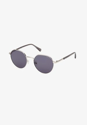 Gafas de sol con lentes redondos y oscuros, marco de metal plateado y brazos negros. Presentan un diseño minimalista y almohadillas nasales ajustables.