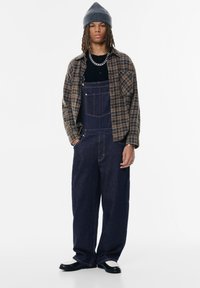 Pettorina in denim di colore blu scuro con tasche laterali, indossata con una maglietta nera e una camicia a quadri marrone. Berretto lavorato a maglia grigio e scarpe a contrasto.