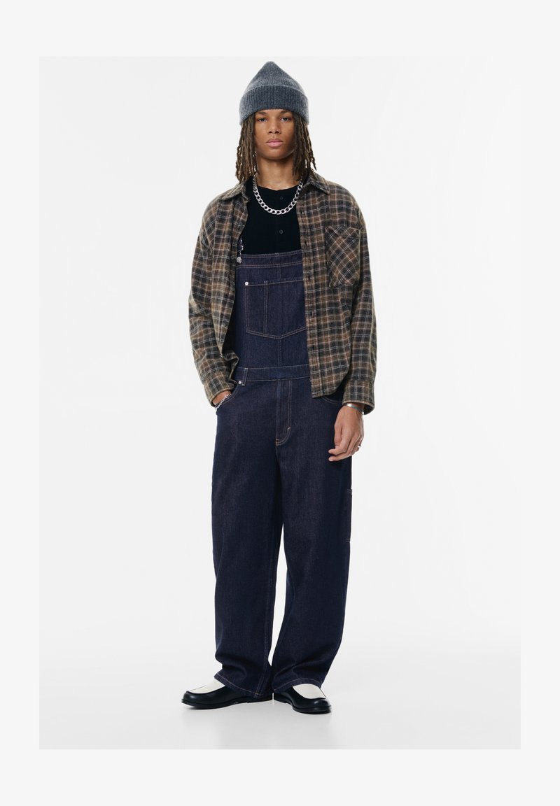 Jeans overal v temno modri barvi s stranskimi žepi, oblečeni z črno majico in rjavo karirano srajco. Sivi pleteni klobuk in kontrastni čevlji.