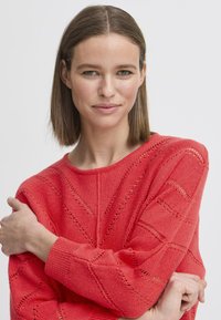 Korallenfarbenes Strickoberteil mit strukturiertem Muster und durchbrochenen Details, runden Ausschnitt und gerippten Bündchen. Weiches, leichtes Material.