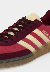 Nærbillede af en rød og burgunderrød Adidas Spezial sneaker med cremefarvede striber, beige tunge, sorte snørebånd og gennemsigtig gummisål.