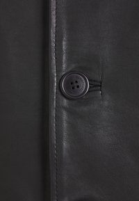 Detalj crne kožne jakne s gumbom s četiri rupe, glatkom teksturom, vidljivim šavovima i elegantnim, sjajnim završetkom.