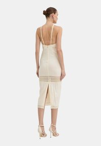 Abito aderente crema con spalline sottili, schiena scoperta e una cerniera. Presenta dettagli in pizzo all'orlo e uno spacco alto fino alla coscia. Indossato con tacchi beige.