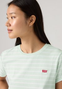 Νεαρή γυναίκα με μακριά σκούρα μαλλιά, που φοράει μικρούς κρίκους στα αυτιά και μια ανοιχτοπράσινη και λευκή ριγέ μπλούζα Levi's, κοιτάζοντας προς τα πλάγια.