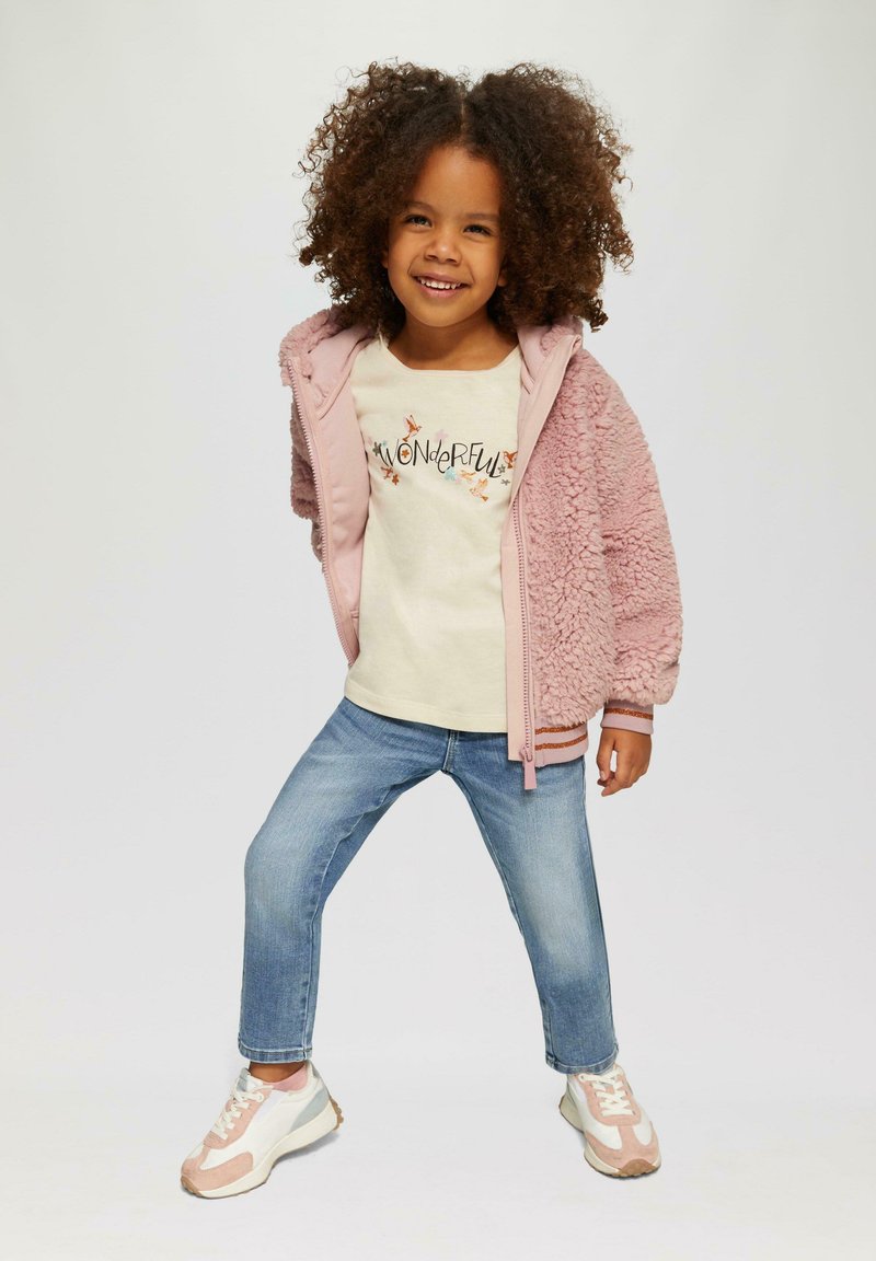 Chaqueta de forro polar rosa claro con capucha, camisa beige con texto "MARAVILLOSO" y acentos florales, vaqueros azules, y zapatillas multicolores.