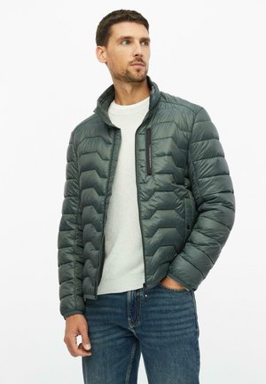 Hombre con una chaqueta acolchada verde oscuro sobre un suéter blanco y vaqueros azules, mirando de lado con una mano en el bolsillo.