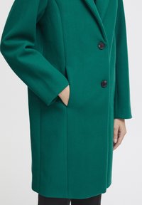 Green wool coat with a structured design, featuring a notched lapel, three black buttons, and side pockets. Folded sleeves and smooth texture.