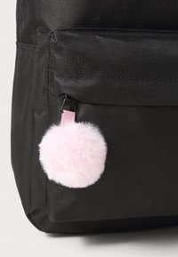 Črn nahrbtnik iz tkanine, ki ima pritrjen roza umetni krzneni pom-pom na zadrgi, prikazuje okroglo obliko in mehak material.