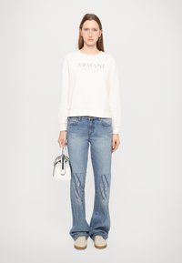 Felpa bianca con logo "Armani Exchange", jeans blu con dettagli logo, sneakers beige e una piccola borsa bianca. Sfondo semplice.