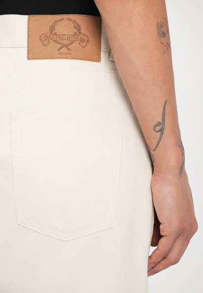 Pantalones de denim color crema con un bolsillo trasero y una etiqueta de cuero marrón que lleva el nombre de la marca "Moschino Milano."