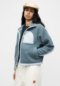 Νεαρό άτομο που φοράει ανοιχτό μπλε φλις μπουφάν, μπεζ καπέλο και λευκό παντελόνι με λογότυπα, στέκεται μπροστά σε απλό λευκό φόντο.