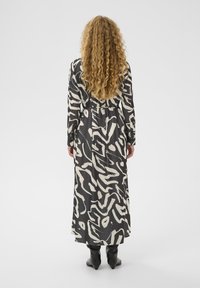 Langes schwarzes Kleid mit einem weißen abstrakten Muster, aus glattem Stoff gefertigt. Verfügt über lange Ärmel und eine taillierte Taille, kombiniert mit schwarzen Stiefeln.