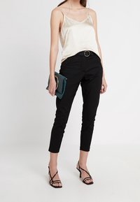 Černé skinny kalhoty v kombinaci s krémovou saténovou topem, černým opaskem s kulatou sponou a tyrkysovou psaníčkem. Outfit doplňují černé sandály na vysokém podpatku.