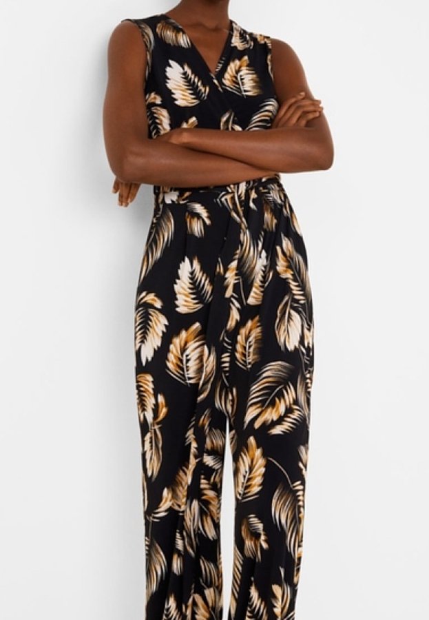 Femme les bras croisés portant une combinaison noire sans manches avec des motifs de feuilles beige et blanc et une ceinture nouée à la taille.