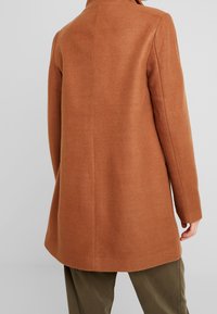 Bruine wollen blendjas met een getailleerde pasvorm, lange mouwen en een gestroomlijnde silhouette. Soepele textuur met een rugventilatie voor meer bewegingsvrijheid.