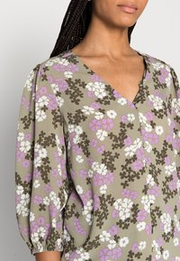 Blusa floral em tecido verde-azeitona, com decote em V, mangas bufantes e padrão floral multicolorido com acentos em roxo, branco e castanho.