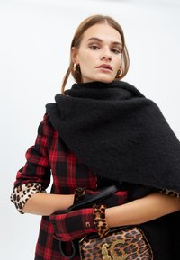 Una modella posa indossando un cappotto a scacchi rossi e un scialle nero, con guanti a stampa leopardata e tiene in mano una borsa a stampa leopardata.