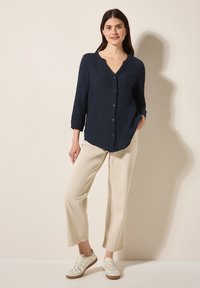 Camicia button-up blu scuro con tessuto testurizzato, maniche a tre quarti e collo rotondo, abbinata a pantaloni beige a gamba larga e sneakers.
