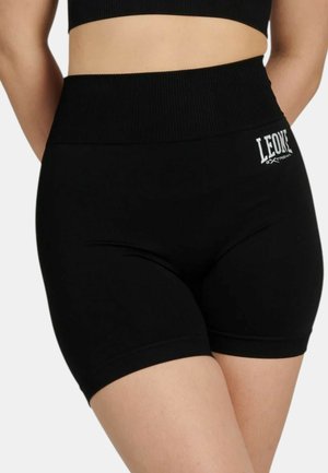 Primo piano di una persona che indossa pantaloncini sportivi neri a vita alta con il logo "LEONE EXTREMA" sull'anca sinistra.