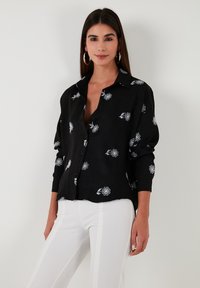 Black button-up shirt made of textured fabric, featuring white floral embroidery throughout and long sleeves. Paired with light-colored pants.