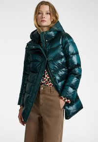 Tyrkysová péřová bunda s vysokým límcem a kapucí, se zlatými kovovými prvky, zipem vpředu a prošívaným designem, doplněná vzorovaným topem a béžovými kalhotami.