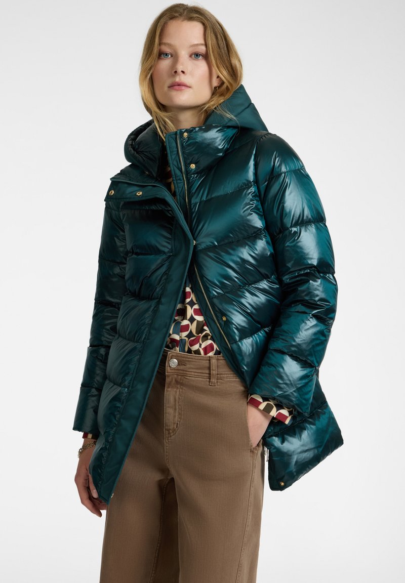 Tyrkysová péřová bunda s vysokým límcem a kapucí, se zlatými kovovými prvky, zipem vpředu a prošívaným designem, doplněná vzorovaným topem a béžovými kalhotami.