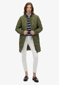 Cappotto lungo trapuntato verde con colletto rialzato, chiusura a zip e polsini a costine, indossato sopra una camicia a righe blu navale e pantaloni bianchi.