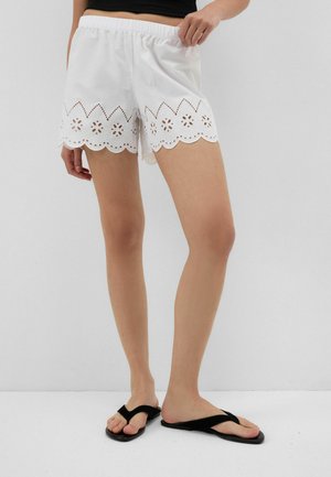 Vrouw draagt witte short met oogjesborduursel en zwarte teenslippers, staand tegen een effen lichtgrijze achtergrond.