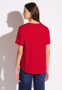 Top corto rosso a maniche corte realizzato in tessuto morbido, con scollatura rotonda e orlo curvo. Il modello indossa jeans scuri.